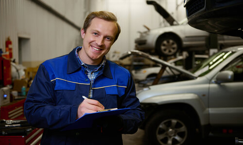 Young caucasian technician smiles looking at camera making checklist for car repair in garage. Car service, repair, car warranty service concept, auto mechanic writes on a clipboard in a workshop.